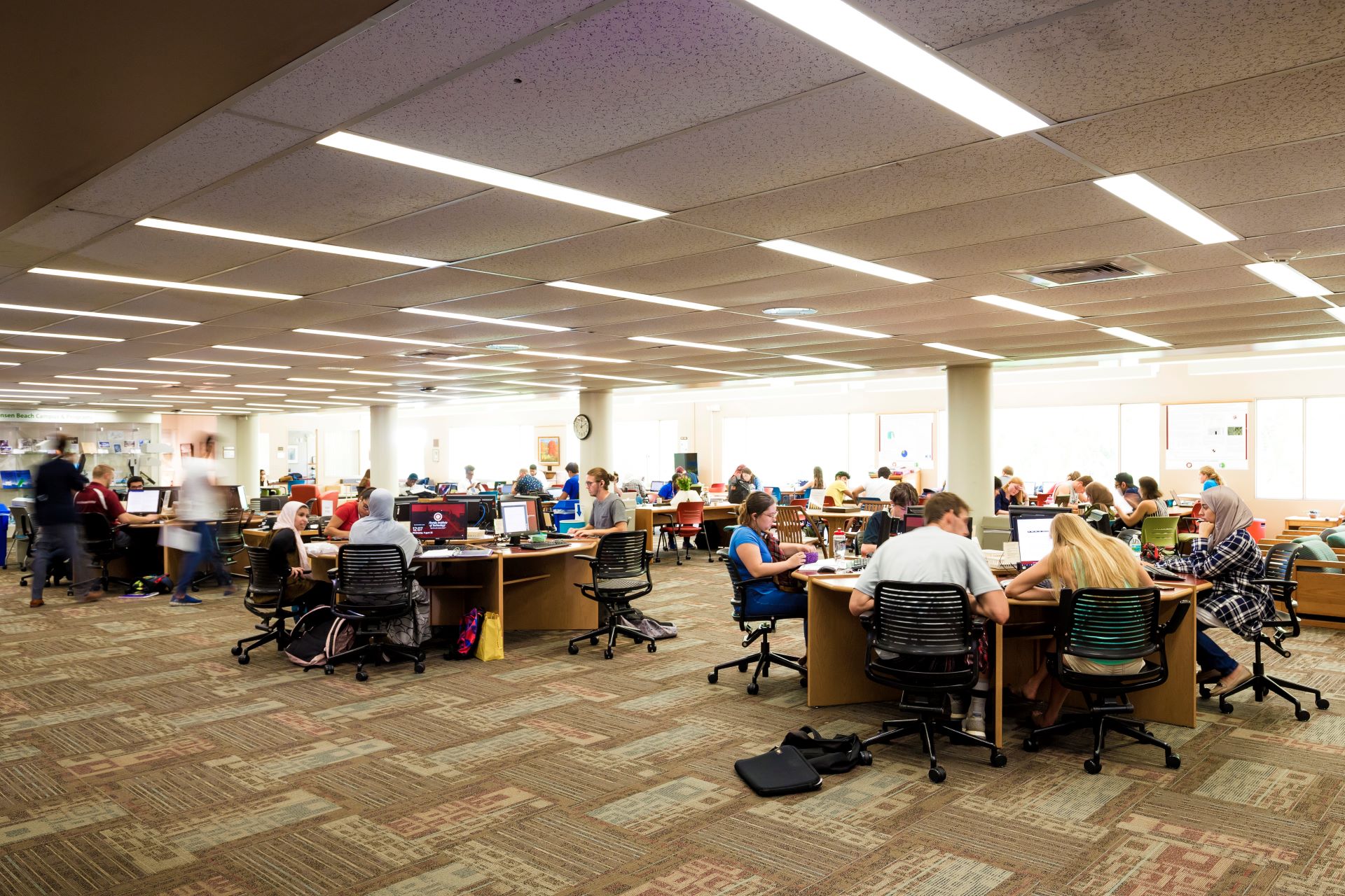 Busy university library with students studying at desks, illuminated by overhead fluorescent lights